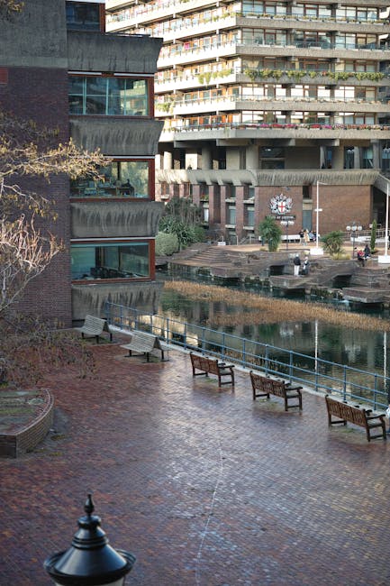 A paved urban courtyard adjacent to modern residential buildings featuring curved balconies and large glass windows. The area includes a narrow waterway with reflective water reflecting the buildings,