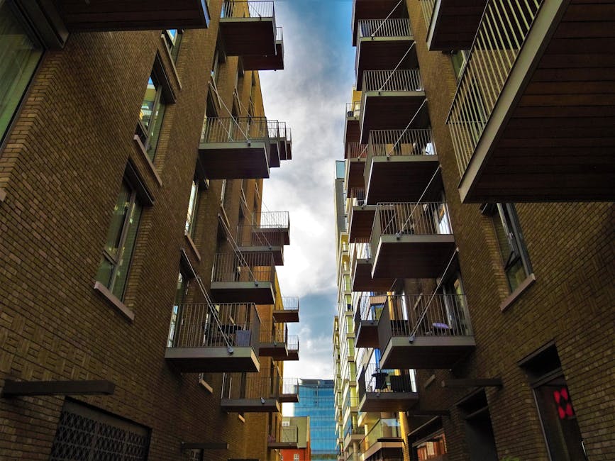 A paved urban courtyard adjacent to modern residential buildings featuring curved balconies and large glass windows. The area includes a narrow waterway with reflective water reflecting the buildings,